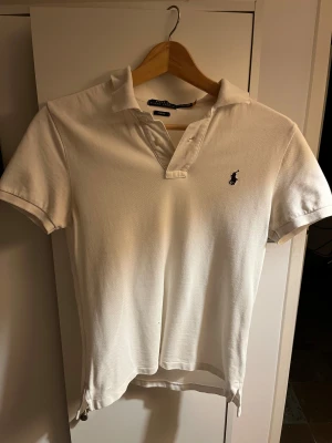 Vit slim fit polo från Ralph Lauren - Vit kortärmad polo från Ralph Lauren i slim fit-modell. Klassisk krage med två knappar och den ikoniska broderade loggan på bröstet. Tillverkad i mjuk bomull som ger en skön känsla och snygg passform. Perfekt för dig som gillar stilrena och tidlösa plagg. Den har dock ett litet hål i sig, se bild