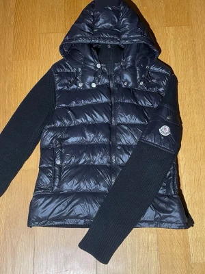 Svart Moncler pufferjacka med huva - Snygg svart pufferjacka från Moncler med stickade ärmar och huva. Jackan har quiltad framsida i glansigt material, dragkedja och Moncler-logga på ärmen. Perfekt för dig som vill ha en cool och varm jacka med exklusiv känsla. den har en liten skada på armen fast märks inte, skriv för mer frågor😁 det är en storlek M fast sitter som en XS