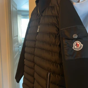 Svart dunjacka från Moncler. - Pris kan diskuteras. Snygg svart dunjacka från Moncler med dragkedja och broderad logga på ärmen. Jackan har en quiltad design och långa ärmar, perfekt för kyligare dagar. Den är både stilren och funktionell med praktiska fickor. Osäker på om den är äkta därför  det nedsatta priset. Priset kan sänkas vid en snabba affär
