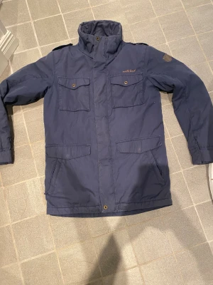 Blå parkajacka från North Bend - Parkajacka i mörkblå färg från North Bend med hög krage och fyra fickor framtill. Jackan har långa ärmar, knappar och dragkedja samt axelklaffar. Materialet är slitstark polyester, perfekt för kyliga dagar. Stl S men passar M 