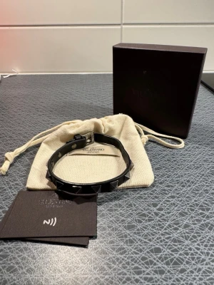 Valentino svart läderarmband med nitar - Säljer ett svart armband från Valentino Garavani i äkta läder med ikoniska pyramidformade nitar i silver. Kommer med originalpåse och box. Perfekt accessoar för dig som vill sticka ut.