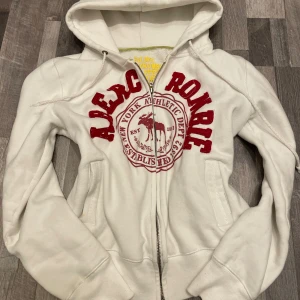 Vit hoodie Abercrombie & Fitch - Säljer en vit hoodie från Abercrombie & Fitch med röd broderad logga och emblem på bröstet. Tröjan har huva med snörning, två fickor fram och dragkedja. Materialet är mjuk bomull och polyester, perfekt för chill dagar. S-M