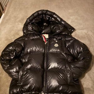 Moncler montbeliard jacka  - Svart pufferjacka från Moncler med glansig finish, huva och dragkedja framtill. Jackan har Moncler-logga på bröstet. Perfekt för kalla dagar och har en riktigt schysst passform. Retail på dessa ligger på runt 21 000kr. Priset går även att diskuteras!