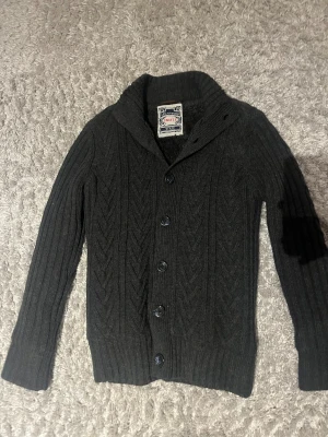 stickad kofta - Säljer en svart stickad kofta från Cedarwood i storlek Small. Koftan har snyggt mönster med diskreta sicksack-ränder, ribbade muddar och knappar framtill. Sitter mer som m/s