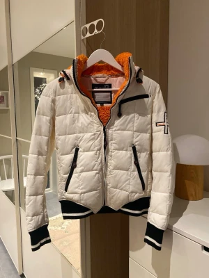 Jacka från Pelle P - Vit jacka från Pelle P med orange teddyfoder, svarta detaljer och dragkedjor. Jackan har ribbade muddar med vita ränder, huva och flera praktiska fickor. Perfekt för dig som gillar sportig och snygg stil.