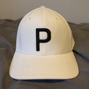 Vit Puma keps med svart P-logga, golfkeps - Snygg vit keps från Puma med stor svart P-bokstav framtill och klassisk justerbar snapback baktill. Kepsen har broderad Puma-logga bak och är tillverkad i ett slätt, lätt material. Perfekt för dig som vill ha en clean och sportig look på golfbanan