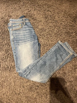 Slim Glenn ljusblå jeans storlek 28/30 - Slim Glenn jeans från Jack & Jones i ljusblå tvätt med snygga slitningar och klassiska fem fickor. Modellen är slim fit med smal passform och raka ben. Jeansen har orangea sömmar och är tillverkade i mjukt bomullsmaterial.