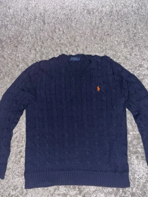 Marinblå stickad tröja Polo Ralph Lauren - Säljer en marinblå stickad tröja från Polo Ralph Lauren med klassiskt kabelstickat mönster och orange broderad logga på bröstet. Tröjan har rund hals, ribbade muddar och är långärmad. Perfekt för dig som gillar stilren och tidlös design.