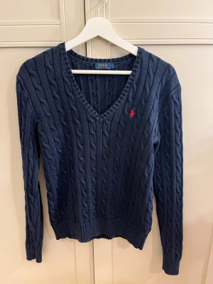 Marinblå stickad tröja Polo Ralph Lauren - Säljer en marinblå stickad tröja från Polo Ralph Lauren med klassisk kabelstickning och röd broderad logga på bröstet. Tröjan har v-ringad hals och långa ärmar. Skulle säga den sitter mera som en M. 