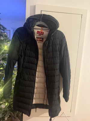 Svart lång dunjacka Parajumpers S - Medellång svart dunjacka från Parajumpers med huva och quiltad design. Jackan har dragkedja framtill, diskret logga på ärmen och ljus insida. Finns ett litet hål ovanför ena fickan, syns knappt och går säkert att laga. 