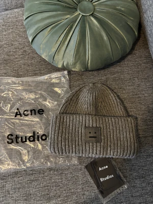 Grå ribbstickad mössa Acne Studios - Säljer en grå ribbstickad mössa från Acne Studios med det ikoniska ansikts-emblemet framtill. Mössan är tillverkad i 100% ull och har en bred uppvikt kant. Perfekt för dig som gillar stilrena accessoarer med en twist.