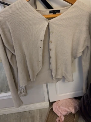 Beige ribbad kofta från Vero Moda - Snygg beige kofta från Vero Moda med ribbad struktur och lång ärm. Koftan har knappar framtill och små håldetaljer på sidorna som ger en chill vibe. Perfekt att slänga över ett linne eller t-shirt för en enkel och clean look.