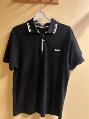 Svart polotopp från BOSS med dragkedja - Svart polotopp från BOSS med vit logga på bröstet och dragkedja framtill. Kragen har snygga vita och beige ränder som ger en fräsch kontrast. T-shirten är kortärmad och har en klassisk, stilren look som funkar till många outfits.