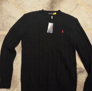 Svart stickad tröja Polo Ralph Lauren - Svart kabelstickad tröja från Polo Ralph Lauren med klassisk rund hals och röd broderad logga på bröstet. Tröjan har långa ärmar och ribbade muddar. Perfekt för dig som gillar stilrena och tidlösa plagg med premiumkänsla.