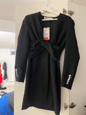 Svart kort klänning från Zara - Svart kort klänning från Zara med snygg omlott-detalj framtill och markerad midja. Långärmad med silvriga knappar vid ärmsluten och djup v-ringning. Perfekt för dig som gillar stilrena och trendiga plagg med en twist.