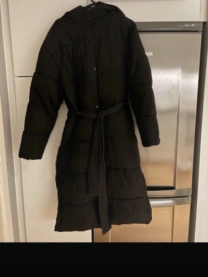 Svart lång pufferjacka med huva - Svart lång pufferjacka med huva och bälte i midjan. Jackan har quiltad design, långa ärmar och knäppning framtill. Perfekt för kalla dagar när du vill hålla dig varm och ändå se chill ut. Enkel och stilren look som funkar till de flesta outfits.