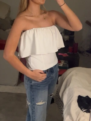 Vit bandeau tubtopp med volang - Säljer en vit bandeau tubtopp med bred volang upptill. Toppen är axelbandslös och har en snygg, figurnära passform. Perfekt att styla med jeans eller kjol för en fräsch look. Materialet känns mjukt och lätt mot huden.