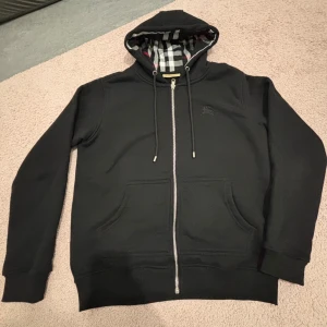 Svart Burberry kofta - Svart Burberry med hel dragkedja, snörning i huvan och klassiskt rutigt foder på insidan. Tröjan har två stora fickor framtill och diskret broderad logga på bröstet. Perfekt för dig som gillar stilrena och exklusiva plagg.