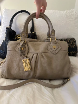 Snygg beige Q Classic Boston bag! - Snyggaste beigea handväskan i äkta läder från Marc by Marc Jacobs. Den ikoniska modellen Q Classic Boston Bag! En populär och eftertraktad vintagemodell med gulddetaljer och dubbla handtag. Väskan är i fint skick, lädret är helt utan repor eller hål. Finns ett märke på undersidan, se bild. Kolla alla bilder och skriv frågor om du är osäker på något!🤍  Det här är en tidlös modell som inte längre finns att köpa, vilket gör den extra eftertraktad. Perfekt för dig som vill ha en stilren tidlös väska