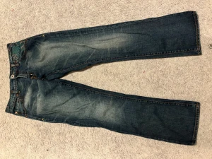 Guess Jeans Premium blå jeans - Snygga blå jeans från Guess Jeans Premium med broderade detaljer i grönt och grått vid fickan. Klassisk femficksmodell med orange kontrastsömmar och dragkedja. Jeansen har rak/utsvänd  passform och är tillverkade i slitstarkt denimtyg.