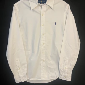 Vit slim fit skjorta från Ralph Lauren - Vit skjorta från Ralph Lauren i slim fit-modell med klassisk krage och långa ärmar. Skjortan har knappar framtill och ett blått broderat Polo-logo på bröstet. Tillverkad i mjuk bomull som känns skön mot huden. Perfekt för dig som gillar stilrena och tidlösa plagg.       Längd 70cm.  Bredd 44cm