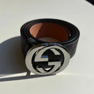 Svart Gucci bälte med GG-spänne - Säljer ett svart Gucci bälte med klassiskt GG-mönster och ett stort, rundat silverfärgat GG-spänne. Bältet är brett och har en lyxig känsla med insida i ljusbrunt skinn. Perfekt för att lyfta din outfit med en ikonisk detalj.