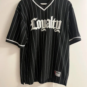 Svart randig fotbollströja Loyalty - Snygg svart fotbollströja med vita tunna ränder och texten 'Loyalty' broderad på bröstet. Tröjan har V-ringning, korta ärmar med vita detaljer och patch nertill med '1992'. Tillverkad i luftig mesh för sportig känsla.