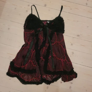 Svart och röd blommig linnetopp med rosett - Säljer en snygg linnetopp med tunna axelband, svart och röd/rosa blommig design och volangdetaljer. Toppen har en söt svart rosett framtill och är gjord i ett lätt, transparent material som känns luftigt. Perfekt för dig som gillar unika detaljer och mönster.