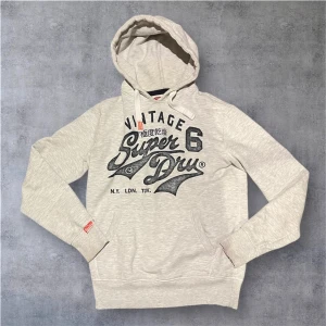 Grå Hoodie från Superdry - Säljer en ljusgrå hoodie från Superdry med svart vintage-tryck på bröstet och coola japanska tecken. Tröjan har huva med snörning, känguruficka och långa ärmar. Perfekt för dig som gillar streetstyle och vill ha något bekvämt och snyggt.