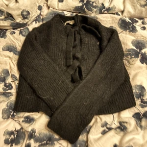 Svart croppad stickad tröja med knyt - Svart croppad stickad tröja med långa ärmar och snygg knytning framtill. Tröjan har en ribbad struktur och är perfekt för lager-på-lager-stil under kyliga dagar. Enkel att matcha med både jeans och kjol. Behövs rengöras ordentligt, därav priset