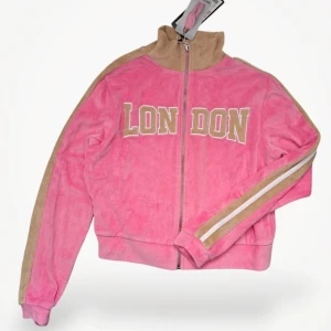Mysig Rosa teddyjacka med LONDON-tryck - Rosa teddyjacka från Sister med beiga detaljer och stort LONDON-tryck framtill. Jackan har dragkedja med ring, hög krage och beigea ränder längs ärmarna. Materialet är mjukt i 80% bomull och 20% polyester. Perfekt för en chill och trendig look..Är oanvänd med alla lappar kvar!