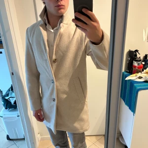Beige rock i ullmix, strl 42 - Snygg beige rock med rak passform och hög krage. Jackan har tre knappar framtill och två sidofickor. Materialet är en mjuk ullmix med polyester och polyamid, vilket gör den både varm och stilren. Perfekt för dig som gillar cleana och klassiska vibes. Bra skick men lätt nopprig. Storlek 42/XS