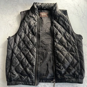 Svart quiltad väst från Oscar Jacobson - Quiltad svart väst från Oscar Jacobson med ståkrage och dragkedja framtill. Västen har ett snyggt rutmönster och elastisk nederkant för skön passform. Perfekt att slänga över en hoodie eller tröja när du vill ha extra värme och stil. Kom gärna med prisförslag!