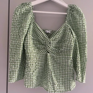Grön och vit rutig blus med twist från mango - Superfin blus i grönt och vitt med rutigt mönster och lång ärm. Toppen har en snygg twist-detalj framtill och smockad rygg för extra komfort. Luftigt bomullsmaterial och fyrkantig ringning ger en trendig vibe. Perfekt till jeans eller kjol för en fräsch look.