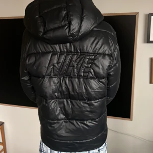 Svart Nike pufferjacka XL barn - Säljer en svart pufferjacka från Nike i storlek 158-170cm med Therma-Fit-teknologi. Jackan har huva, dragkedja framtill, stora fickor och broderad Nike-logga på bröstet och ryggen. Perfekt för kalla dagar och riktigt snygg streetstil.