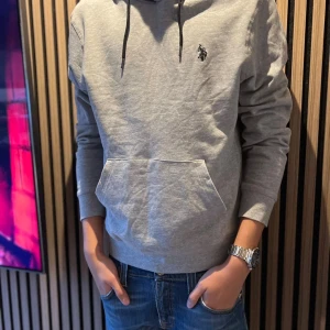 Grå hoodie från U.S. Polo - Snygg grå hoodie från U.S. Polo Assn. med svart logga på bröstet och svarta snören i huvan. Tröjan har en klassisk känguruficka framtill och ribbade muddar vid ärmslut och nederkant. Perfekt för chill dagar och enkel att matcha med jeans.