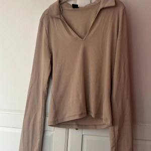 Beige långärmad polotopp - Säljer en beige långärmad polotopp med v-ringning och krage. Toppen har en enkel och clean design, tillverkad i mjukt material som känns skönt mot huden. Perfekt för dig som gillar minimalistisk stil och neutrala färger.