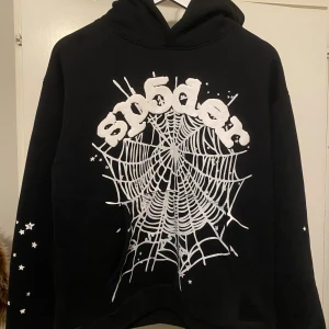 Svart Sp5der hoodie med vit spindelnätsprint - Svart hoodie från Sp5der i storlek S med stor vit spindelnätsprint och logga på framsidan. Ärmarna har vita stjärnor som detaljer. Tröjan har huva och är långärmad, perfekt för dig som gillar streetwear med unik design.