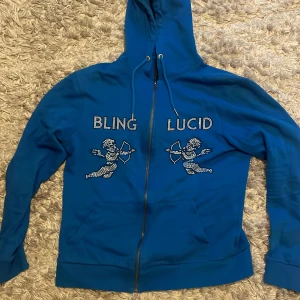Blå hoodie med bling och ängelmotiv - Snygg blå hoodie från Bling Lucid med glittrande stenar på bröstet och coola ängelmotiv. Tröjan har huva med snörning, dragkedja framtill och rymliga fickor. Perfekt för dig som gillar statement-plagg och vill sticka ut med lite extra bling.