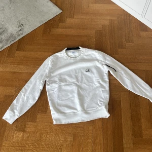 Vit sweatshirt från C.P. Company - Vit sweatshirt från C.P. Company med broderad logga på bröstet. Klassisk rund halsringning, ribbade muddar och långärmad modell. Tillverkad i mjuk bomull som är skön att bära. Perfekt för en clean och stilren look.