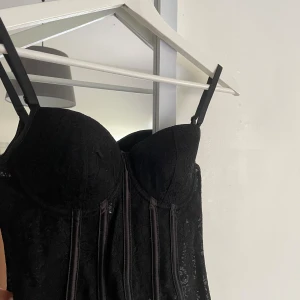 Svart spetskorsett med snörning - Säljer en svart korsett med justerbara axelband och snygga spetsdetaljer. Toppen har markerade kupor och dekorativa sömmar framtill. Baksidan har snörning för att kunna anpassa passformen. Perfekt för dig som vill ha en edgy och trendig look.