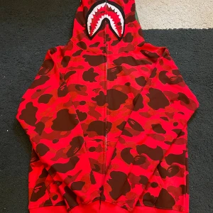 Röd camo hoodie från BAPE Shark - Säljer en ikonisk röd camo hoodie från BAPE med det klassiska haj-motivet på huvan. Hoodien har hel dragkedja, huva och coolt kamouflagemönster i rött och brunt. Perfekt statement-plagg för streetwear-stilen.