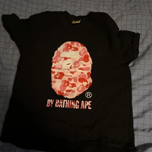 BAPE t-shirt  - Svart t-shirt från BAPE med ikoniskt ape-head tryck i rosa och röd camo på bröstet samt texten 'BY BATHING APE' under. Klassisk passform och korta ärmar. Tillverkad i Japan, riktigt streetwear-vibe.