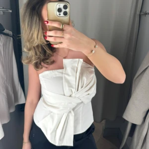 Vit omlott tubtopp med knyt - Säljer en snygg vit tubtopp med omlott-design och stor knytning framtill. Toppen är axelbandslös och har en cool, strukturerad form som ger en unik look. Perfekt för dig som vill sticka ut med en stilren och modern topp. Dragkedja baktill