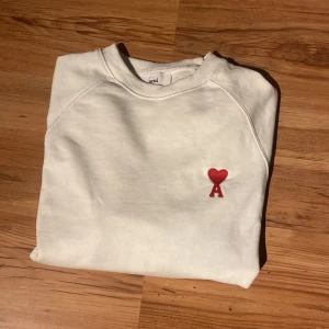 Vit sweatshirt från Ami med rött hjärta - Vit sweatshirt från Ami med klassisk passform och rund hals. På bröstet finns ett broderat rött hjärta med bokstaven A. Tröjan har långa ärmar och ribbade muddar vid ärmslut och nederkant. Perfekt för en clean och stilren look.