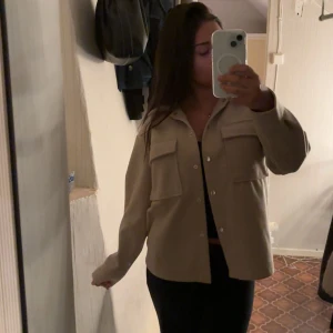 Beige vårjacka från Gina Tricot - En beige vårjacka från Gina Tricot med stora större fickor och knappar framtill. Skjortan har en rak passform och långa ärmar. Storlek Xxs men är större i passformen och inte kort i ärmarna, jag brukar bära storlek S i jackor