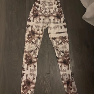 Stronger leggings med brun batikmönster - Snygga leggings från Stronger med unikt batikmönster i bruna och beige toner. Hög midja och stretchigt material som sitter skönt på kroppen. Perfekta för träning eller när du vill ha en cool look. Logga på höften och diskreta sömmar för extra stil. Använda några få gånger💗
