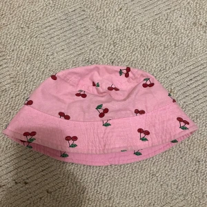 Rosa bucket hat med körsbärsmönster - Söt rosa bucket hat med tryckta körsbär över hela hatten. Hatten har en klassisk form med nedvikt brätte och är tillverkad i ett mjukt bomullstyg. Perfekt accessoar för soliga dagar och ger en lekfull touch till din outfit.