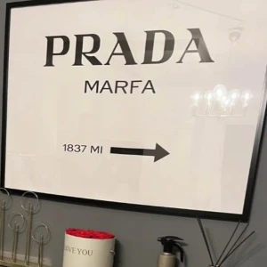 PRADA MARFA - Stilren poster med texten 'PRADA MARFA' och en pil som visar avståndet 1837 miles. Perfekt för dig som gillar mode och vill ge ditt rum en trendig och modern känsla. Passar utmärkt som inredningsdetalj för unga vuxna och modeintresserade. Har 2 st, en oanvänd i förpackning och en som hänger. Den förpackade skickas. 