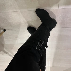 Knähöga stövlar helt nya, storlek 36.5-37.5 - Säljer ett par helt nya svarta höga stövlar med tre snygga spännen på utsidan. De har platt sula och rund tå, tillverkade i mocka-look som ger en stilren vibe. Perfekta för att lyfta din outfit under kallare månader.  Amerikansk skostorlek 5.5 (eu 36.5-37)